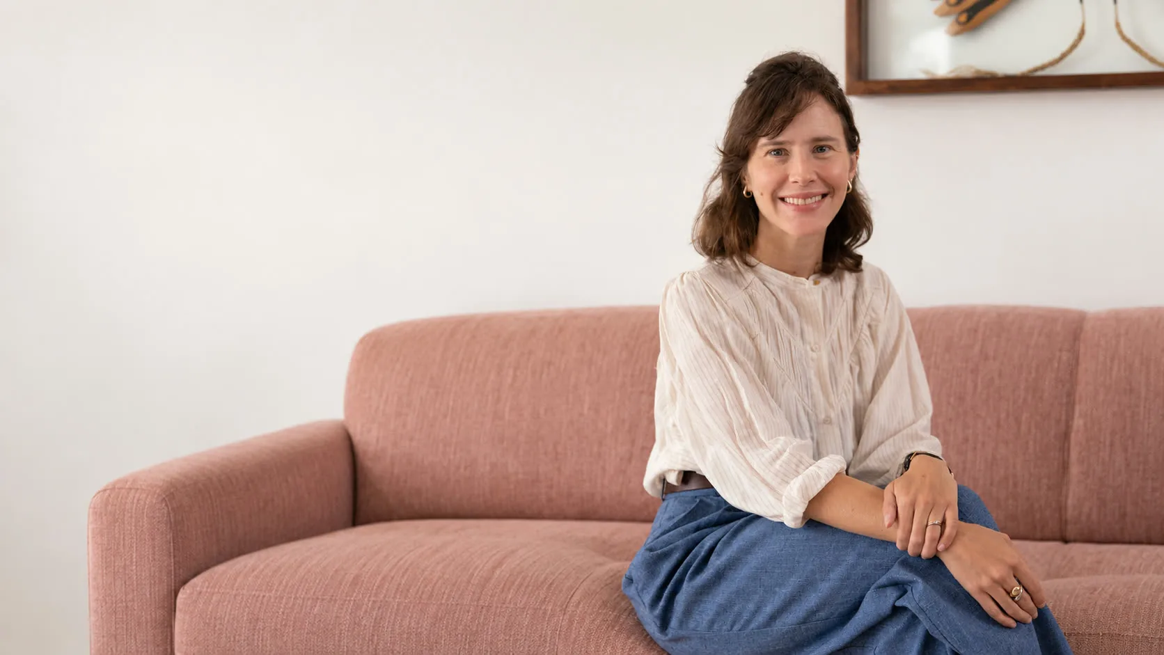 Renata Camargo sorrindo, de blusa branca, sentada num sofá rosado